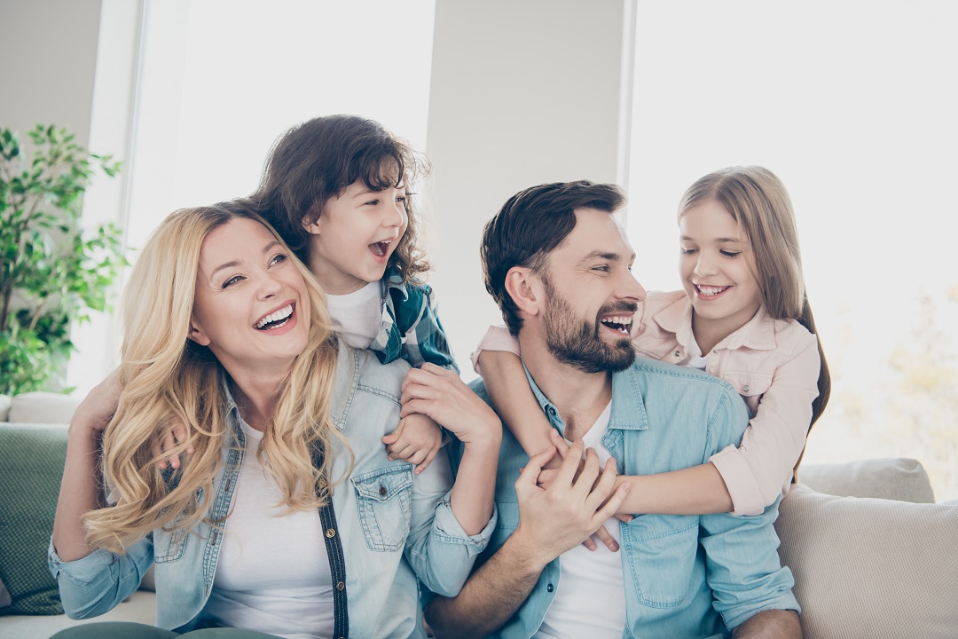 A family of four poses together on a couch, with the father holding a child and smiling at the camera.