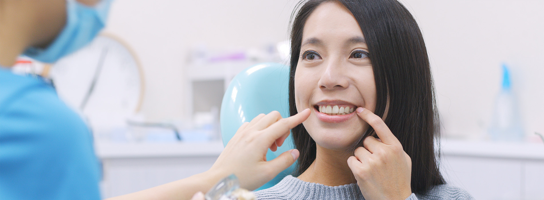 The image shows a woman sitting in a dental chair with a smile on her face, being attended to by a dental professional who appears to be adjusting or cleaning her teeth.