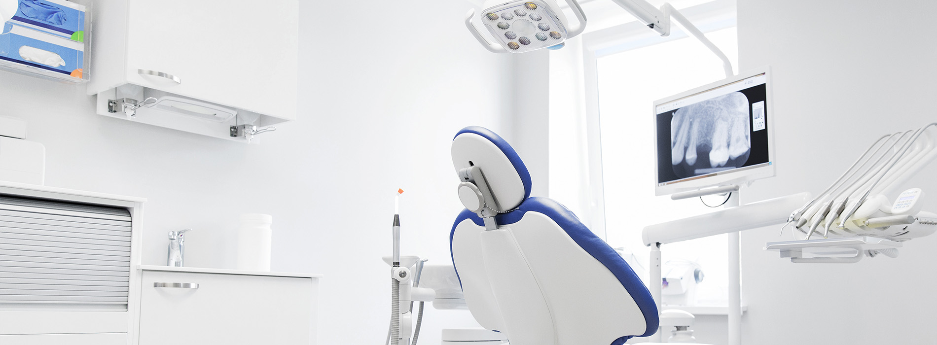 The image depicts a modern dental office interior with a dental chair and equipment, including a blue dental chair and various medical instruments.