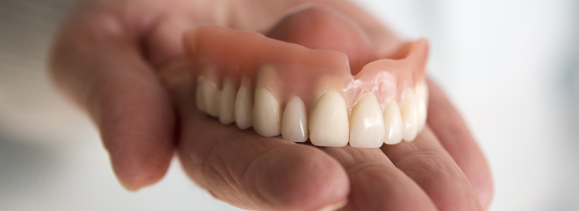 The image shows a person s hand holding an upper denture with artificial teeth against a blurred background.