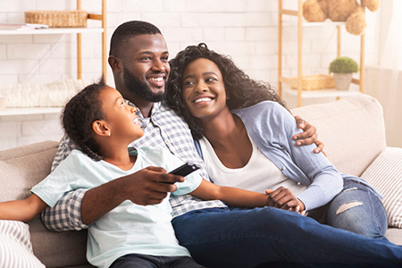 A family of four sitting together on a couch, with two adults and two children, smiling and looking at something off-camera.