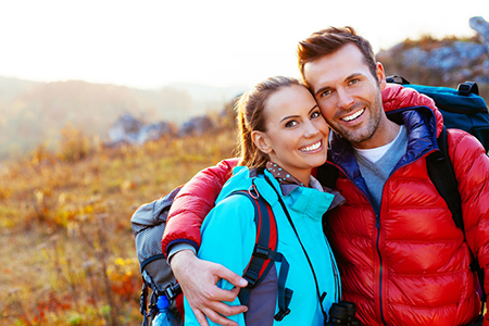 A man and woman are embracing outdoors, both wearing backpacks and smiling, with a scenic landscape behind them.