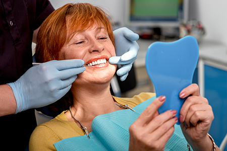The image shows a woman sitting in a dental chair with her mouth open, receiving dental care from a professional who is holding a mirror up to the patient s face.
