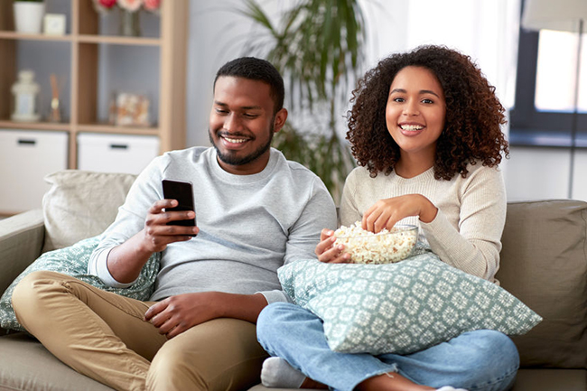 A man and woman sitting on a couch, smiling at each other while enjoying popcorn, with the man holding a smartphone.